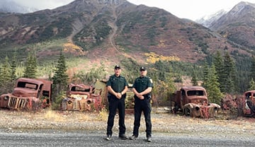 2 conservation officers stand in front of a row of abandoned Second World War vehicles.