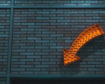 An orange arrow sign with small lightbulbs in front of a dark grey wall