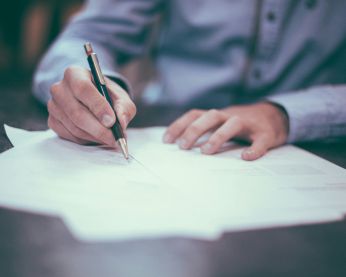 Person filling out a form - image shows a pile of papers with a person writing on the top page