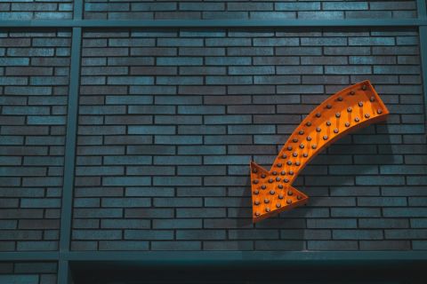 An orange arrow sign with small lightbulbs in front of a dark grey wall