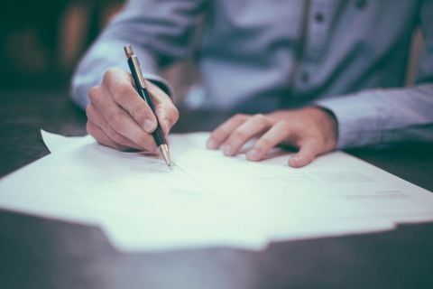 Person filling out a form - image shows a pile of papers with a person writing on the top page