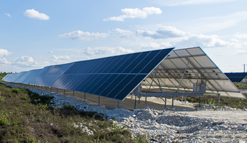 A row of solar panels in a field with sunny blue sky and scattered clouds above.